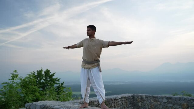 Adult Indian man rising form a hatha yoga pose doing a stretch at sunrise on castle walls outside in nature in traditional yogi clothes