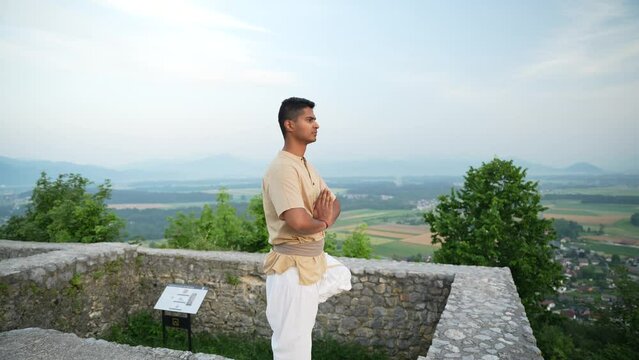 concentrated open-eyed Indian man doing a hatha yoga pose standing on one leg overlooking the valley from a top of a hill at sunrise at dawn