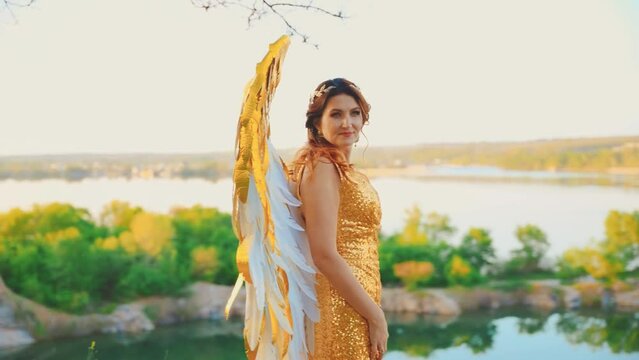 Fantasy woman goddess angel golden white wings long gold dress standing on top of mountain looking back turns. view on river magic divine blue sky. Art Fairy girl sexy lady creative image costume 4k