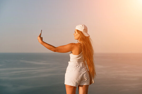 Selfie woman sea. The picture depicts a woman in a cap and tank top, taking a selfie shot with her mobile phone, showcasing her happy and carefree vacation mood against the beautiful sea background