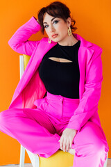 a woman in a bright suit sits on a chair posing on an orange background