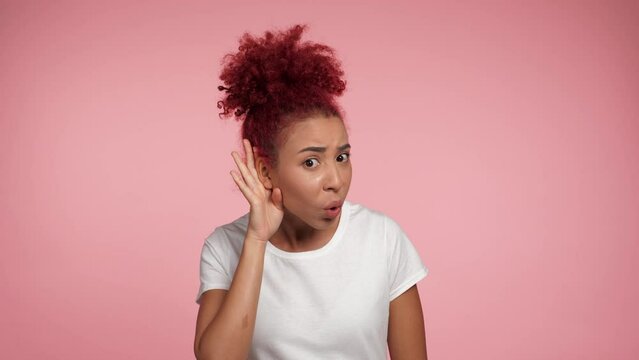 Portrait confused African American redhead curly woman with hand near ear, cannot hear what is being said. Close up female wearing white t-shirt standing on isolated pink background with copy space