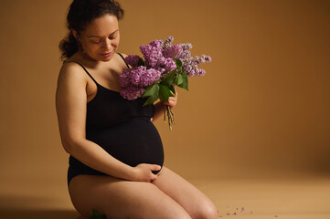 Beautiful multi-ethnic adult pregnant woman in black top, stroking her belly, expressing happy positive emotions while feeling baby kicks, holding blooming lilac flowers, isolated on beige background