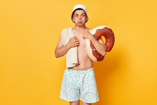 Funny attractive man wearing shorts swimsuit and panama holding donut rubber ring isolated on yellow background holding breath blowing cheeks.