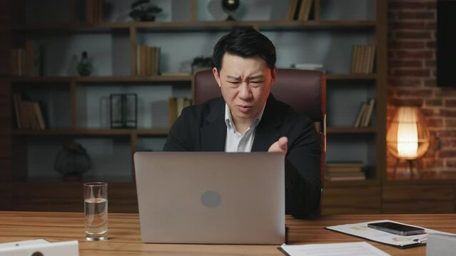 Middle aged asian male worker looking at laptop screen and being shocked of gadget breakdown or operational problems. Man spreading hands in confusion and nervously exclaiming what.