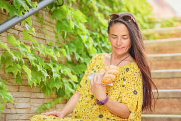 Obraz premium Positive attractive brunette woman in yellow dress sitting on stairs with bagel