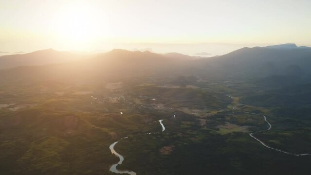 Aerial drone flight over green tropical forest at sunset. Mountain view at golden hours. Narrow river. Green hills landscape. Orange-yellow tone. Exotic nature and travel concept. Calm water surface.