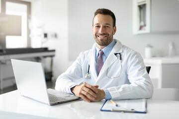 Portrait of man general practitioner working on laptop in medical office, preparing to attend appointment, looking at camera and smiling