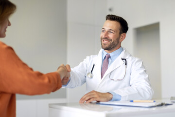 Cheerful male GP shaking hand of female patient at consultation in clinic. Man physician...
