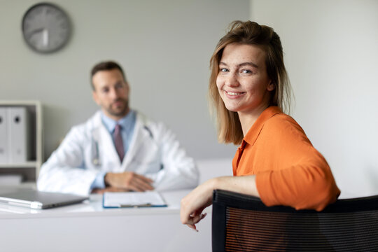 Portrait of happy young female patient feeling satisfied with good quality service in clinic, sitting at consultation with male doctor in hospital and smiling at camera