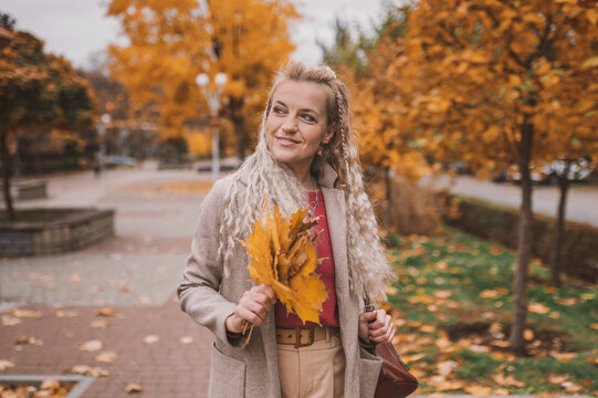 Attractive middle age woman in trendy coat in front of autumn leaves enjoys warm weather. Mature woman walks in the autumn park and enjoys an active life.