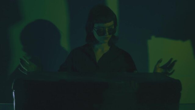 Young woman in black round sunglasses carefully opening a suitcase or a treasure chest. Glowing treasure inside. Dark room. Spy movie, burglar, thief theme. Discovery, treasure concept. Cinematic.