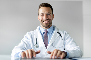 Happy friendly male doctor typing on laptop keyboard, consulting and answering patients questions, working in clinic interior and smiling at camera