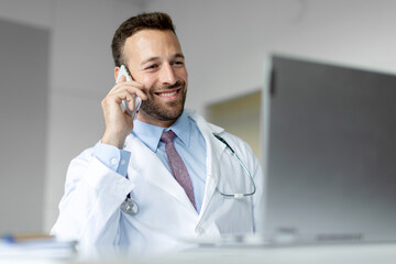 Cheerful male doctor talking on cellphone and using laptop, therapist having mobile conversation with patient, giving recommendations online - Powered by Adobe