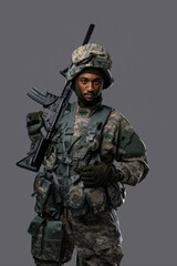 Confident soldier in NATO uniform and helmet poses with his rifle on a plain grey background, showcasing the courage and strength of military personnel