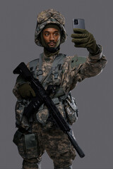 Dark skinned soldier in NATO uniform and helmet holds up his smartphone for a selfie or video chat on a plain grey background, showing the versatility and technology skills of military personnel