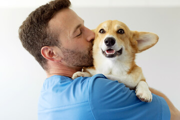 Friendly man veterinarian holding on hands and kissing cute pembroke welsh corgi dog during examination in the clinic, closeup shot - obrazy, fototapety, plakaty