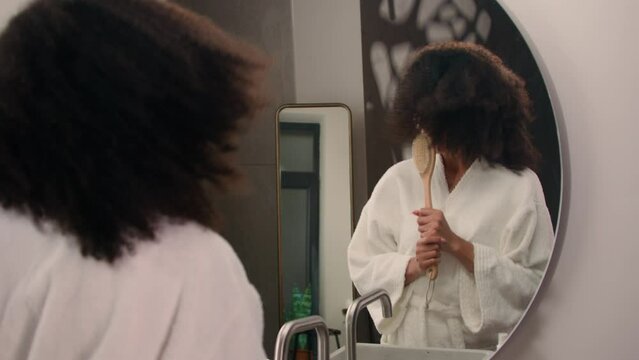 Happy carefree African American woman singing in hairbrush as microphone ethnic girl having fun in bath looking in mirror sing in brush dance pretend singer fooling around beauty routine in bathroom