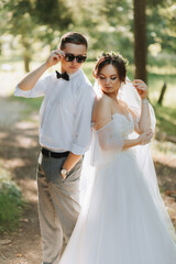 Naklejka premium Fashionable groom and cute bride in white dress with tiara of fresh flowers, hugging, laughing in park, garden, forest outdoors. Wedding photography, portrait of smiling newlyweds.