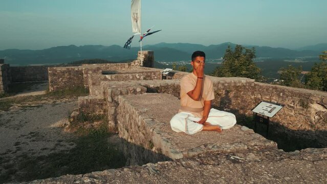 Drone shot of Indian man sitting in hatha yoga pose and meditating nadhi shuddhi on stone castle wall on top of the hill surrounded by hills fields and forests in the morning at sunrise