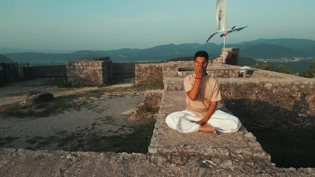 orbit drone shot of Indian man sitting in hatha yoga pose and meditating nadhi shuddhi on stone castle wall on top of the hill in traditional yogi clothes kurta and dhoti at sunrise
