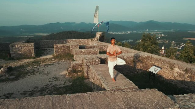 orbit slow motion shot of Indian man doing hatha yoga standing on one leg on stone castle wall on top of the hill surrounded by hills fields and forests in the morning at sunrise