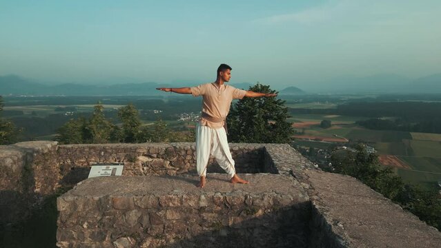 Indian man doing hatha yoga pose at sunrise on stone castle wall on top of the hill barefoot and in traditional meditation clothes with fields and trees behind him