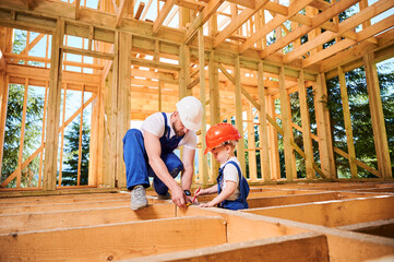 Father with toddler son building wooden frame house. Male worker playing with kid on construction site, wearing helmets and blue overalls on sunny day. Carpentry and family concept.