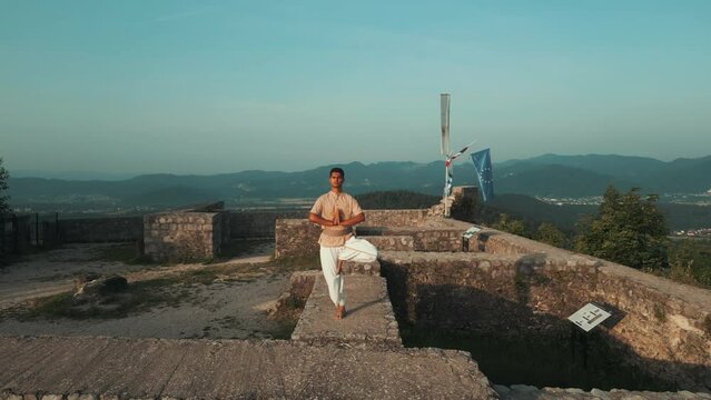 Drone shot of Indian man standing in hatha yoga pose standing on one leg on stone castle wall on top of the hill surrounded by hills fields and forests in the morning at sunrise