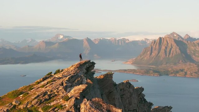 Hiker on arctic waterfront mountain top with glacier view in midnight sun; drone