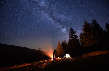Overnight camping expedition, under shimmering stars. Man and woman intrepid hikers near bonfire and tent pitched near woods, under resplendent night sky with Chumack Path as their celestial guide.