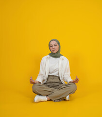 Woman in hijab sitting on the ground, spreading hands in yoga om aum gesture. Relaxing, meditate calm dawn. Isolated yellow background. Closed eyes. Enjoying meditation. Healthy lifestyle concept.