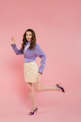 doll pose, beautiful young woman with wavy hair gesturing and looking at camera, trendy outfit, brunette model in purple jacket and high heels posing on pink background, studio shot, conceptual