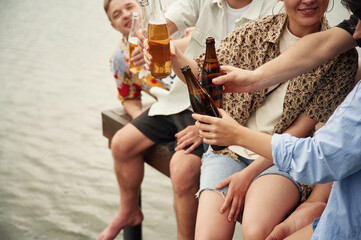 Group of friends are sitting near the lake with beer in hands