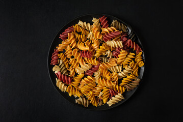 Multi-colored pasta in the form of a spiral on a black plate on a dark background. Photo in low key