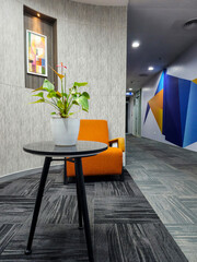 In the modern office area, white flower pot adorned with a laceleaf plant sits elegantly upon a sleek black table, orange chair positioned gracefully behind it.