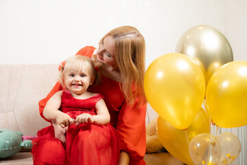 Mother and two-year-old daughter in red dresses sit near balloons