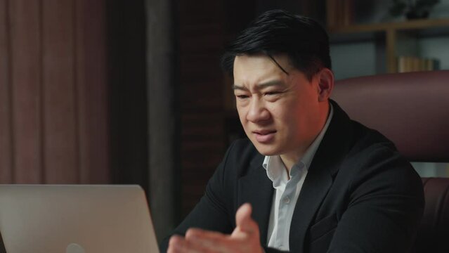 Close up of worried asian boss loading programs on computer and getting errors on screen. Officially dressed entrepreneur spreading hands in annoyance while sitting in upscale office.