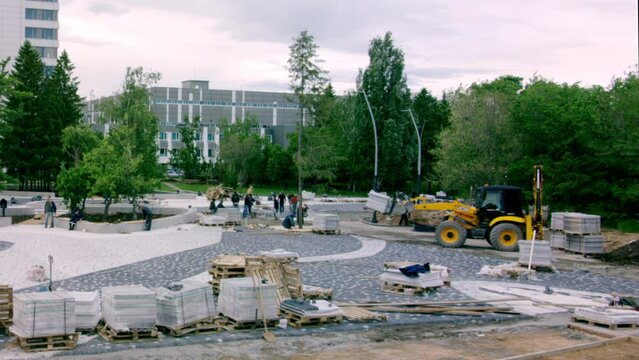 Excavator moving stone slabs for sidewalk pavement around park at daytime. Stone bricks and construction debris scattered around park timelapse