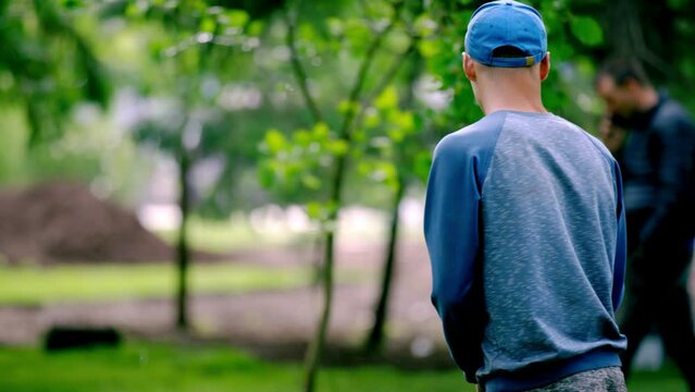 Male employee uses garden hose to provide hydration to trees on park lawn. Man wanders around park during gardening work