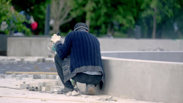 Employee in dirty clothes uses electric cutter to cut bricks in downtown. Worker cuts paving slabs during repair of footpath backside view