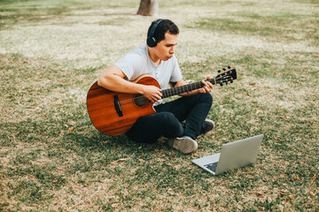 Photograph of young man playing guitar and teaching on a laptop in a park. Concept of people,...