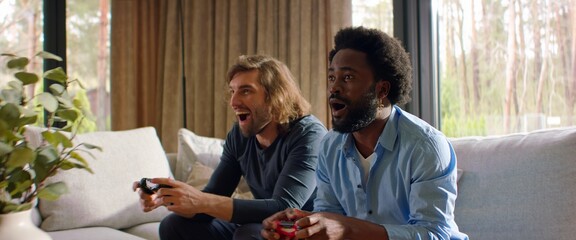 Two 30s male friends - African-American and Caucasian, playing video games at home, laughing and...