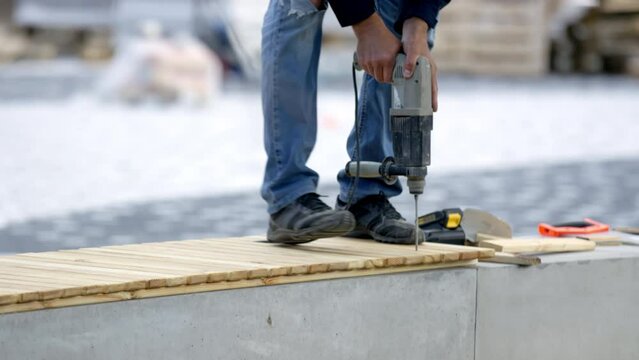 Employee in uniform uses drill to put wood planks together to construct a bench at park renovation. Worker stands on the bench to drill wooden boards at daytime