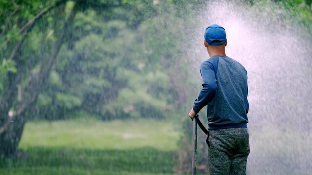 City worker in regular clothing waters green lawn. Man sprays water on grass. Employee uses garden hose to water trees in city park