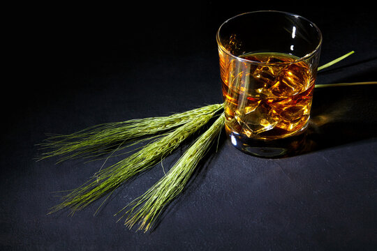 Glass of whiskey with ice in a glass cup surrounded by ears of wheat on a black stone table