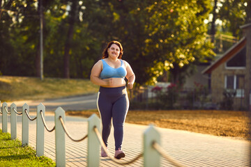 Happy overweight woman jogging in park. Smiling motivated beautiful fat large plump stout lady in sports top bra and yoga pants running on paved park path on sunny morning or evening. Fitness concept