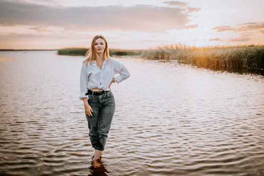A woman in jeans and a white shirt stands in the water at sunset on the shore of a lake