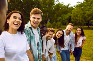 Obraz premium Portrait of friends having fun and laughing while walking in park on warm summer day. Multiracial men and women in casual clothes stand in row behind each other and smile outdoors. Selective focus.