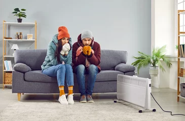 Crédence en verre imprimé Pleine lune Full length photo of a young frozen family couple man and woman sitting on sofa in the living room in winter outerwear and in hats at home warming their hands. Heating problems concept.
